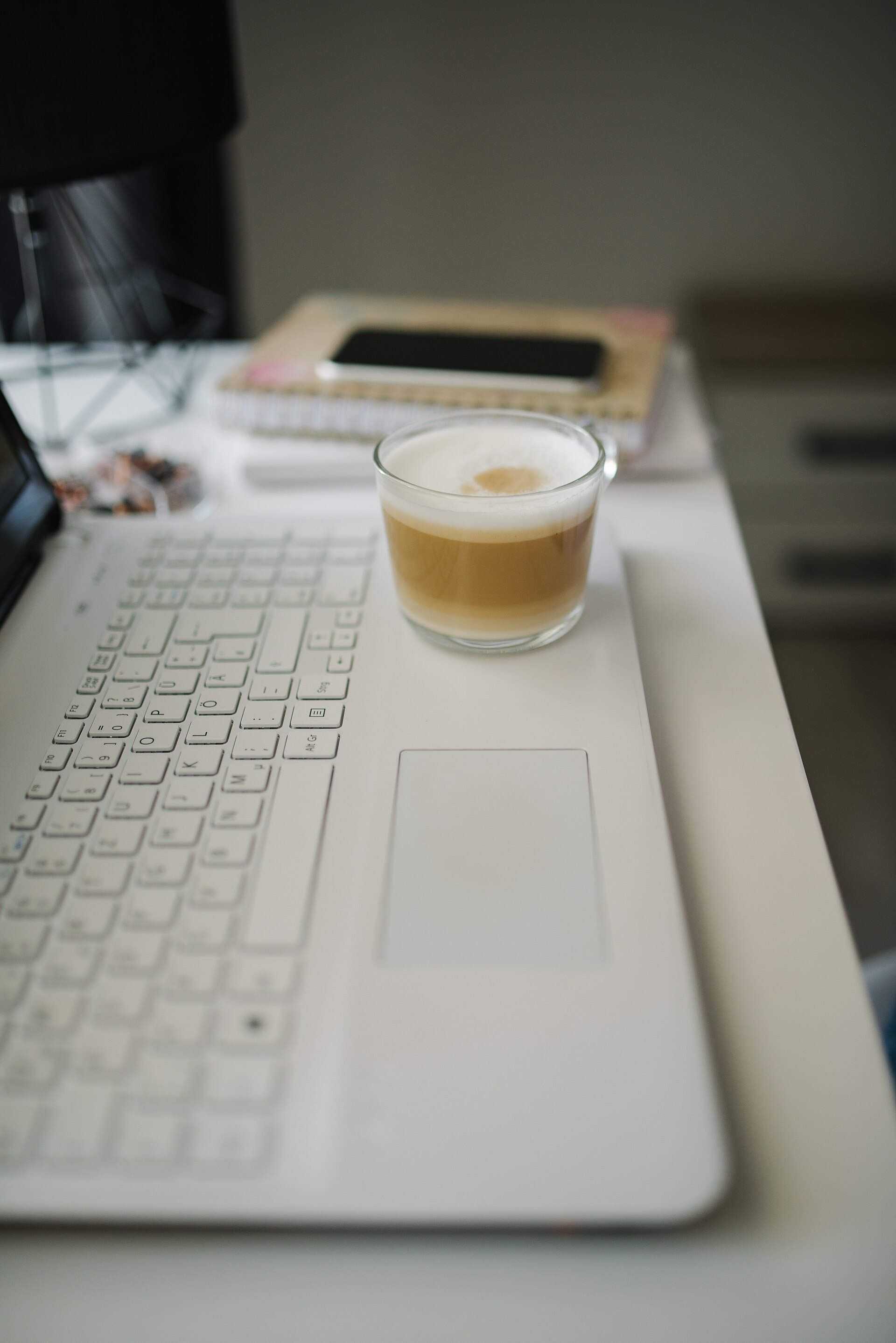 Kaffe ved en bærbar computer med dansk tastatur, notesbog og telefon på skrivebordet
