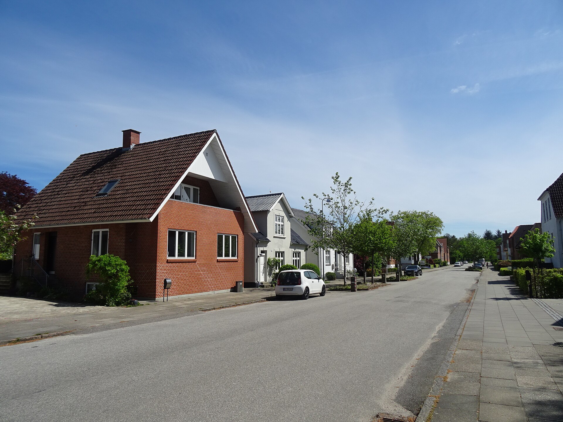 Stille boligvej i Herning med rødstenshuse, grønne træer og en parkeret bil i solskin